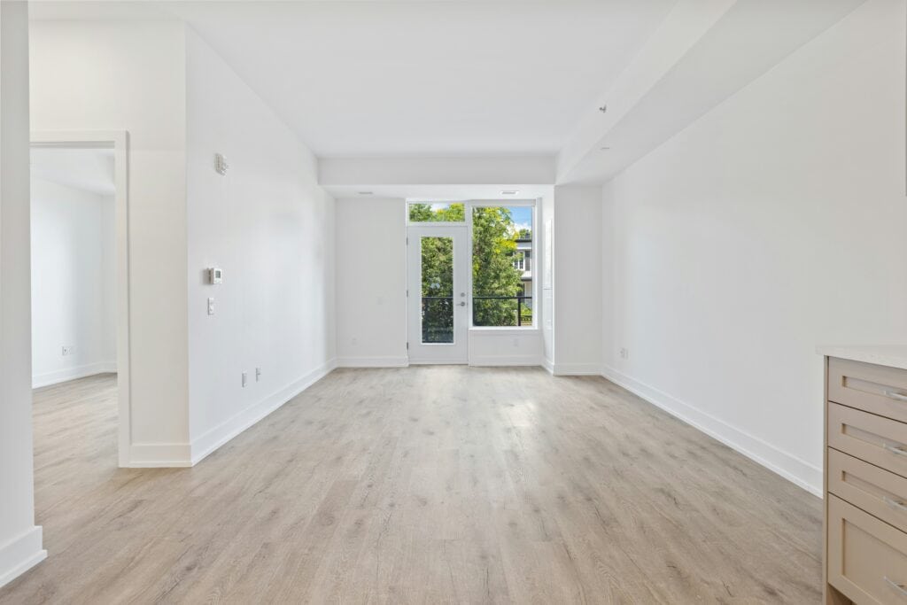 Empty apartment room with pale wood flooring and glazed doors opening onto greenery